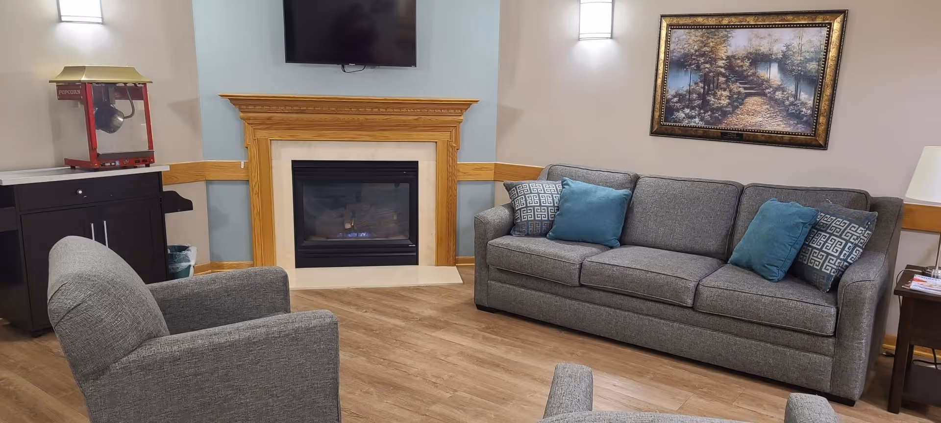 A cozy living room area with a gray sofa and matching armchairs, decorated with blue and patterned throw pillows. There is a wooden fireplace with a TV mounted above it, a popcorn machine on a black cabinet, a framed painting on the wall, and a small side table with a lamp and magazines.