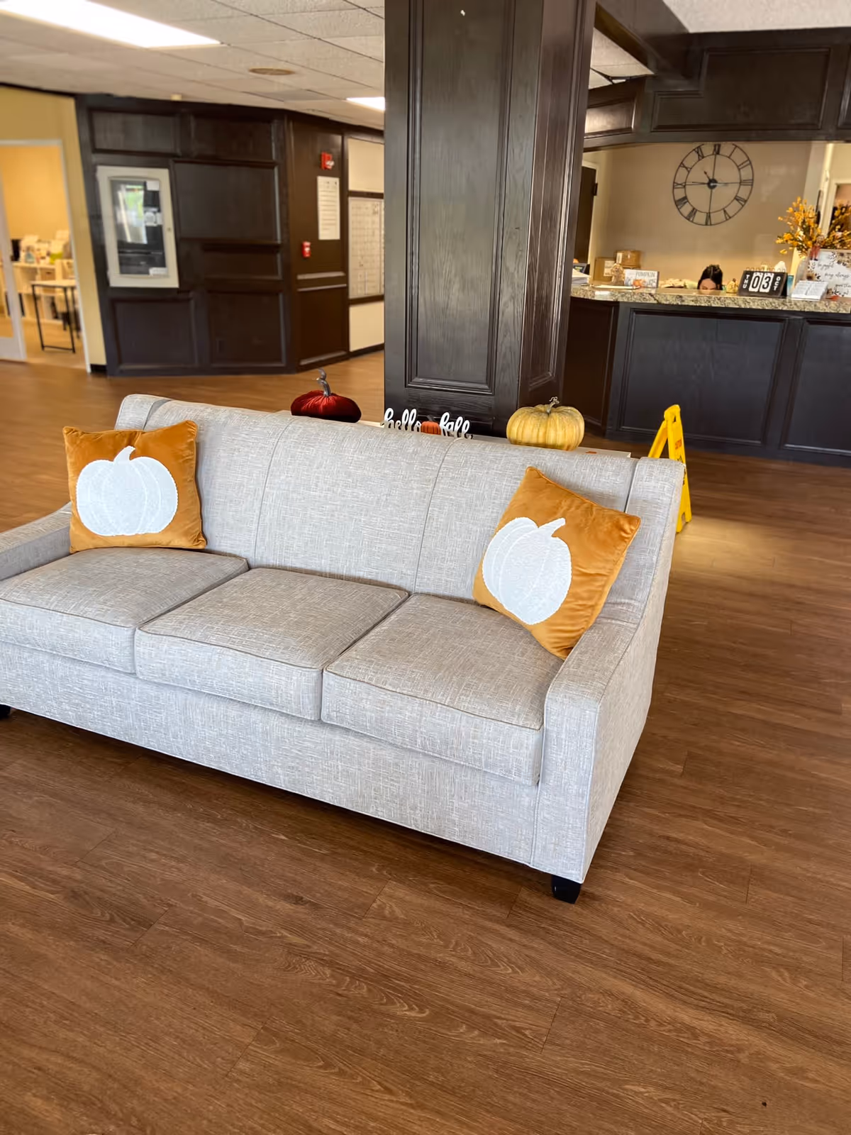 A beige sofa with two orange pillows featuring white pumpkin designs is placed on a wooden floor in a senior living facility lobby. Behind the sofa, there is a dark wooden column decorated with small pumpkins and a 'hello' sign. In the background, a reception desk with a large wall clock and some autumn decorations is visible.