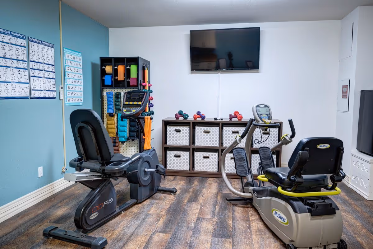 A small fitness room with two recumbent exercise bikes, a wall-mounted TV, a storage unit with baskets and dumbbells, and workout posters on the wall.