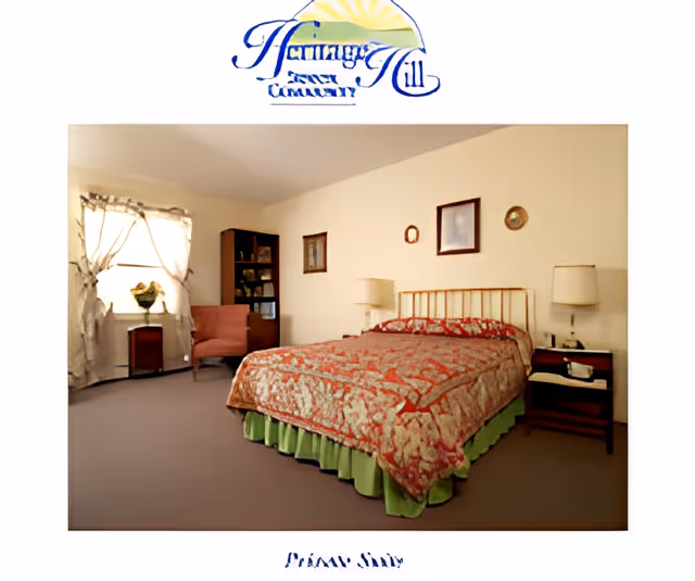 A cozy bedroom in a senior living facility with a double bed covered in a patterned bedspread and green bed skirt. The room has beige walls with framed pictures, two bedside tables with lamps, a window with sheer curtains, a small wooden cabinet, a pink upholstered chair, and a bookshelf filled with books and decorative items.