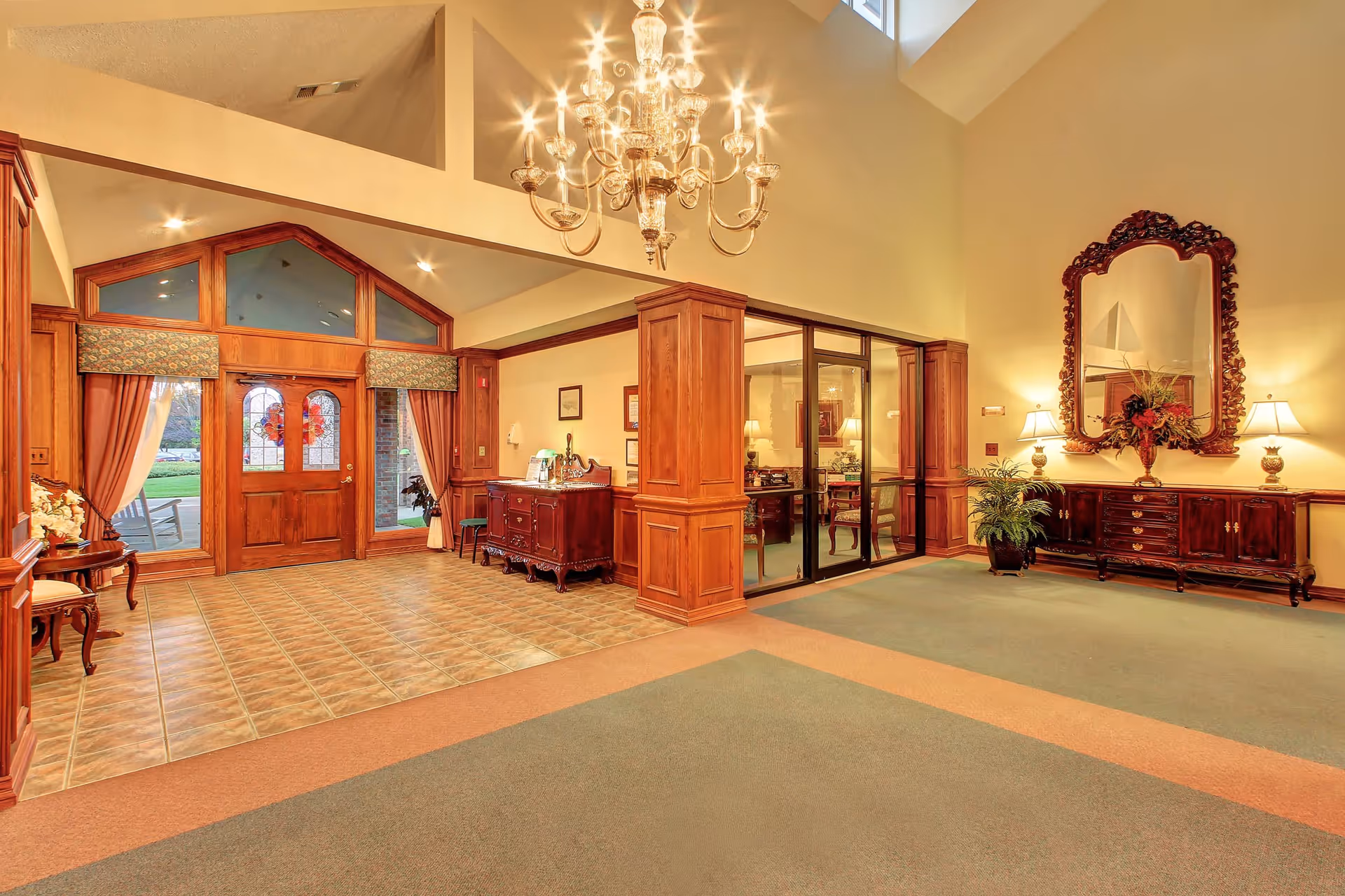 Spacious and elegant interior lobby area with a high ceiling, a large chandelier, wooden double doors with glass panels, and classic wooden furniture including a sideboard with a large ornate mirror and a small table with flowers. The floor is a combination of tiled and carpeted sections, and there are large windows letting in natural light.