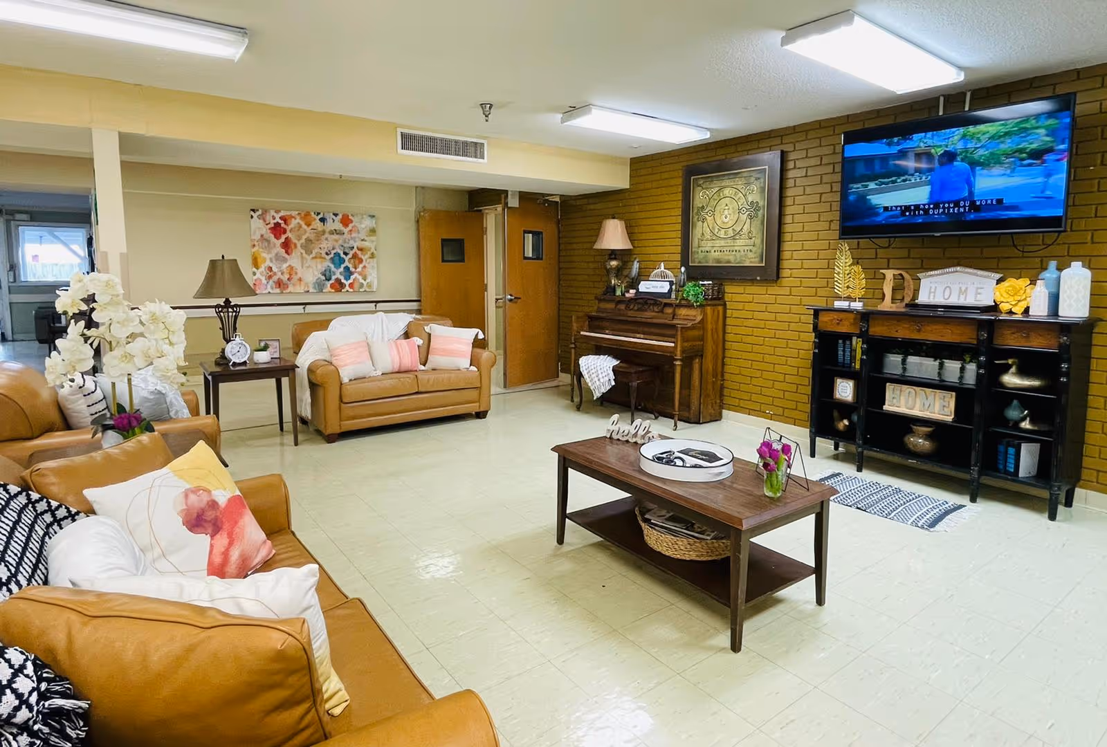 A cozy living room with tan leather sofas adorned with colorful pillows, a wooden coffee table with decorative items including a small vase with flowers, a piano against a brick wall, and a flat-screen TV mounted above a black console table decorated with home-themed signs and vases.