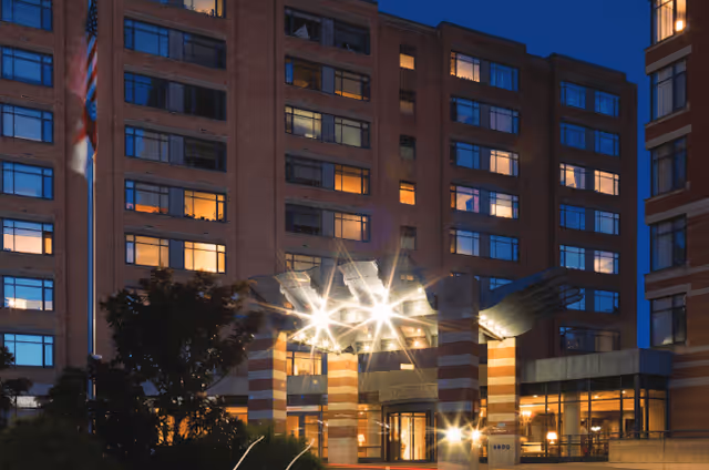 Exterior view of a multi-story senior living facility building at dusk with illuminated windows and entrance. The entrance features a distinctive architectural design with large columns and a sculpture resembling outstretched hands. Trees and landscaping are visible in front of the building.