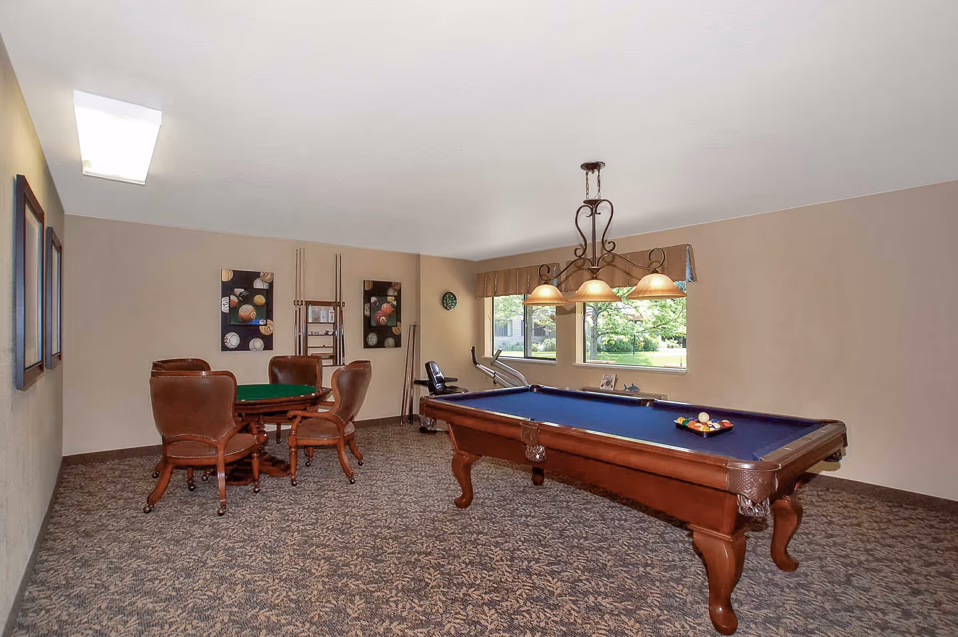 A recreational room with a pool table featuring a blue felt surface and a rack of billiard balls arranged on it. There is a hanging light fixture with three lamps above the pool table. In the background, there is a small round table with four brown leather chairs on casters. The walls are beige with framed pictures of billiard balls and a rack holding pool cues. Two windows let in natural light and show greenery outside. The floor is carpeted with a patterned design.