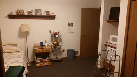 A small room with a single bed on the left side covered with striped bedding. Next to the bed is a wooden nightstand with various items on it, including a lamp and a small shelf above holding framed pictures and decorations. There is a floor lamp beside the nightstand. In the corner, there is a metal rack with flowers and other items. A door is visible on the back wall, and to the right, there is a walker, a microwave on a shelf, and a small refrigerator underneath. The room has carpeted flooring and plain walls.