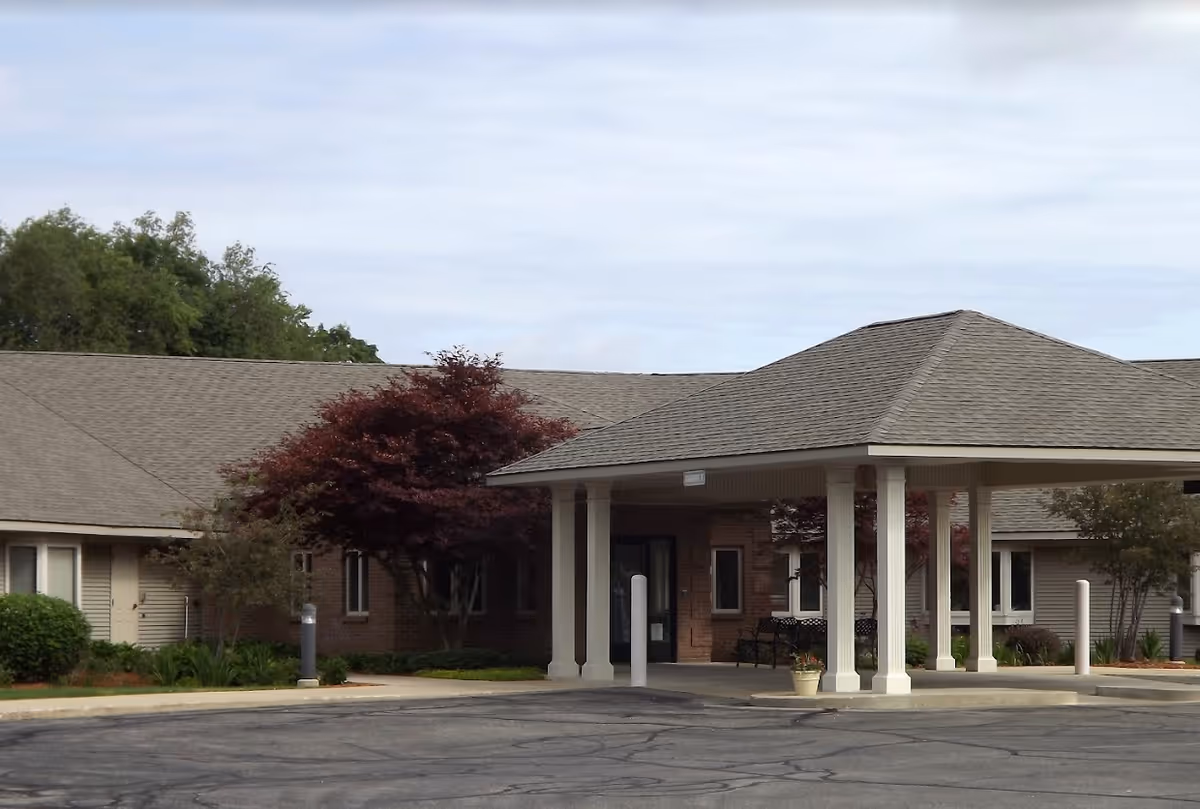Front entrance of a single-story assisted living building with a covered porte-cochere, white columns, and landscaped shrubs.