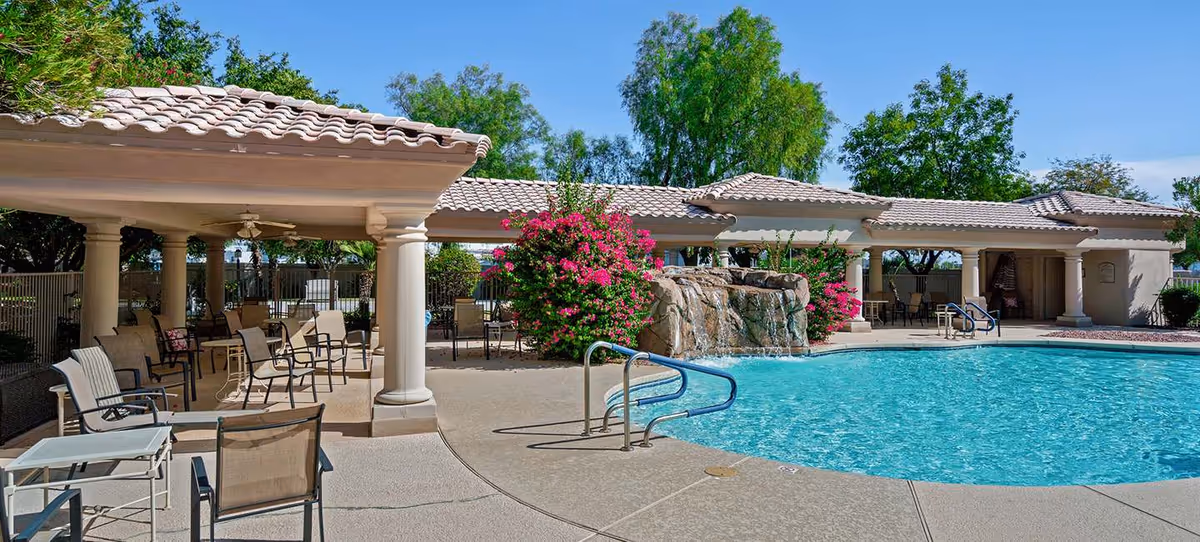 Sunlit outdoor swimming pool with a rock waterfall, covered patio and chairs at a senior living community.