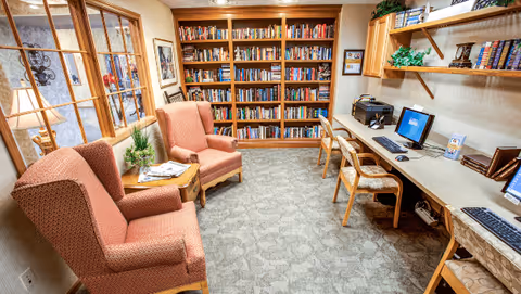 A cozy room with two upholstered armchairs and a small wooden table between them, a large bookshelf filled with books against the back wall, and a long desk with three chairs and computer workstations on the right side. The room has carpeted flooring and warm lighting.