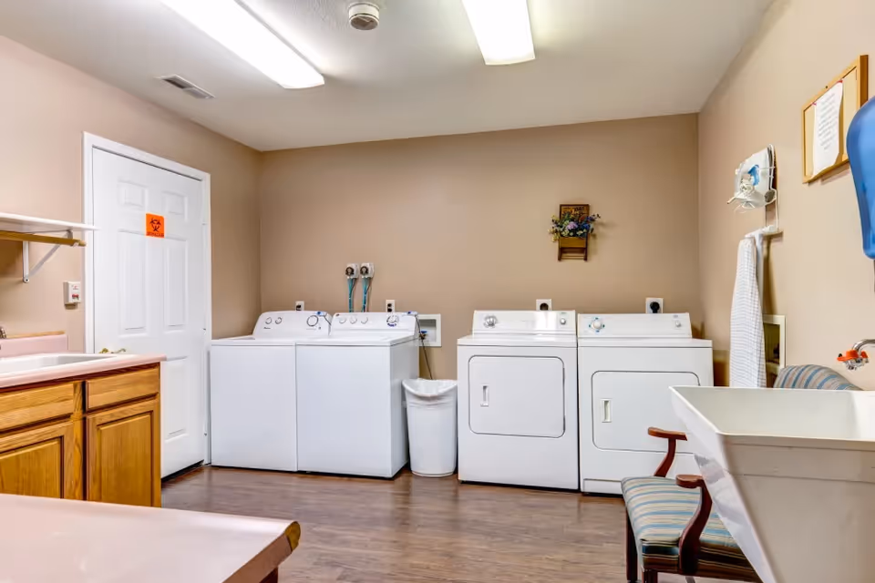 Laundry room with two washing machines and two dryers against a beige wall. There is a white trash bin between the washers and dryers. A wooden cabinet with a pink countertop is on the left side, and a utility sink with a striped chair is on the right. The room has wood flooring and fluorescent ceiling lights.