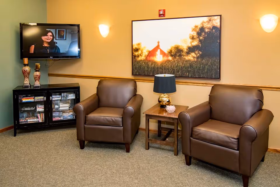 A cozy sitting area with two brown leather armchairs separated by a wooden side table with a black and gold lamp and a small decorative bowl. On the wall behind the chairs is a large framed picture of a barn at sunset. To the left, a flat-screen TV is mounted on the wall above a black cabinet filled with books and DVDs. The walls are painted in warm tones with two wall sconces providing soft lighting.