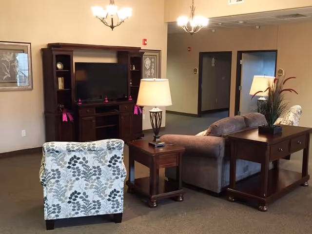 A cozy living room area in a senior living facility with a floral patterned armchair, a gray sofa, wooden side tables with lamps, a wooden entertainment center with a TV, and framed artwork on the walls. The room is softly lit by ceiling chandeliers and has a neutral color palette.