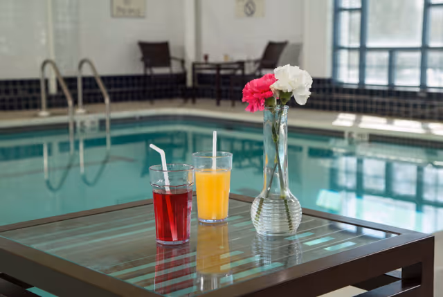 Two glasses with straws, one filled with a red drink and the other with an orange drink, placed on a glass-top table next to a vase with pink and white flowers. In the background, there is an indoor swimming pool with chairs and tables along the poolside.