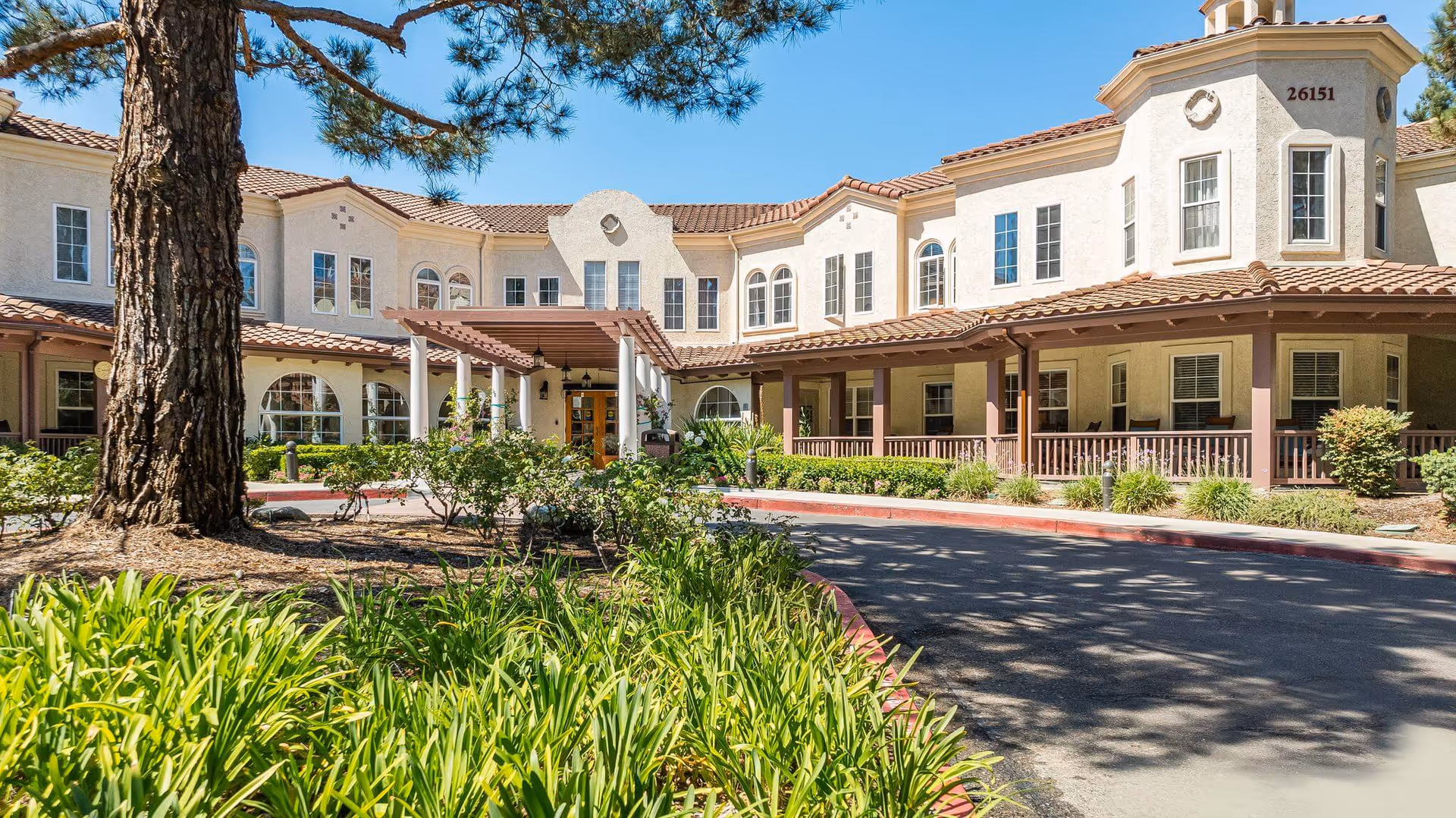 Front exterior of a two-story Mediterranean-style senior living building with a covered entrance, driveway, and landscaped grounds.