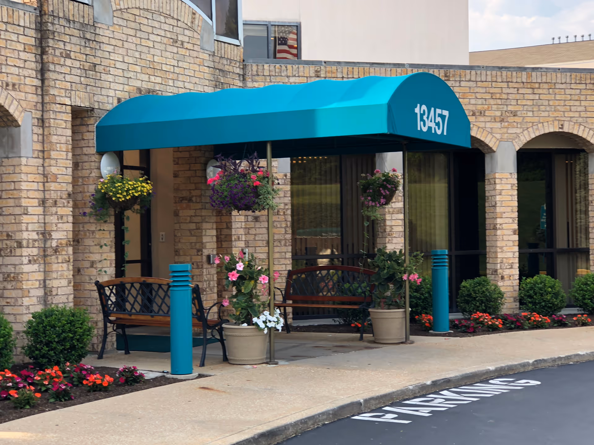 Entrance of a building with a teal awning displaying the number 13457. The entrance is framed by brick walls and has two benches under the awning. There are hanging flower pots and potted plants with colorful flowers around the entrance. The sidewalk and part of the parking lot with a 'PARKING' marking are visible.