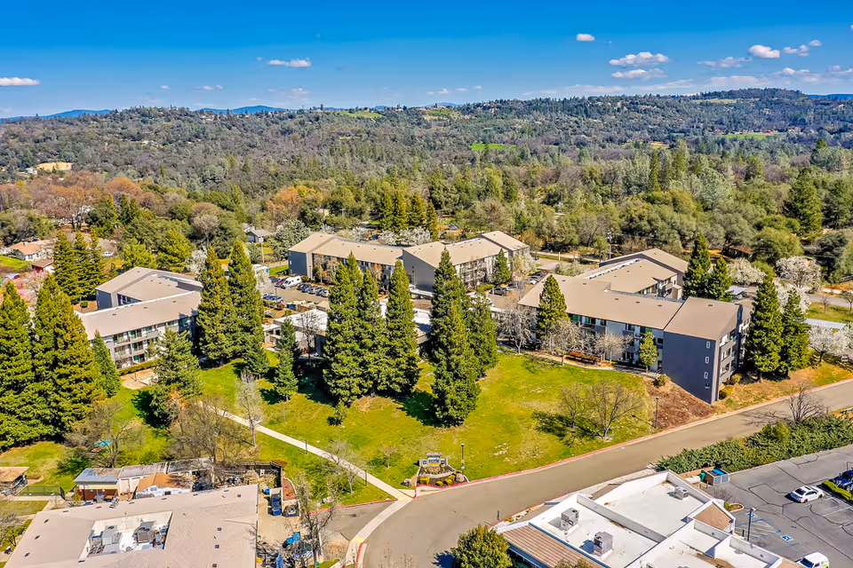 Aerial view of the Gold Country Retirement Center complex showing multiple low-rise buildings, open lawns and tall trees set against wooded hills.