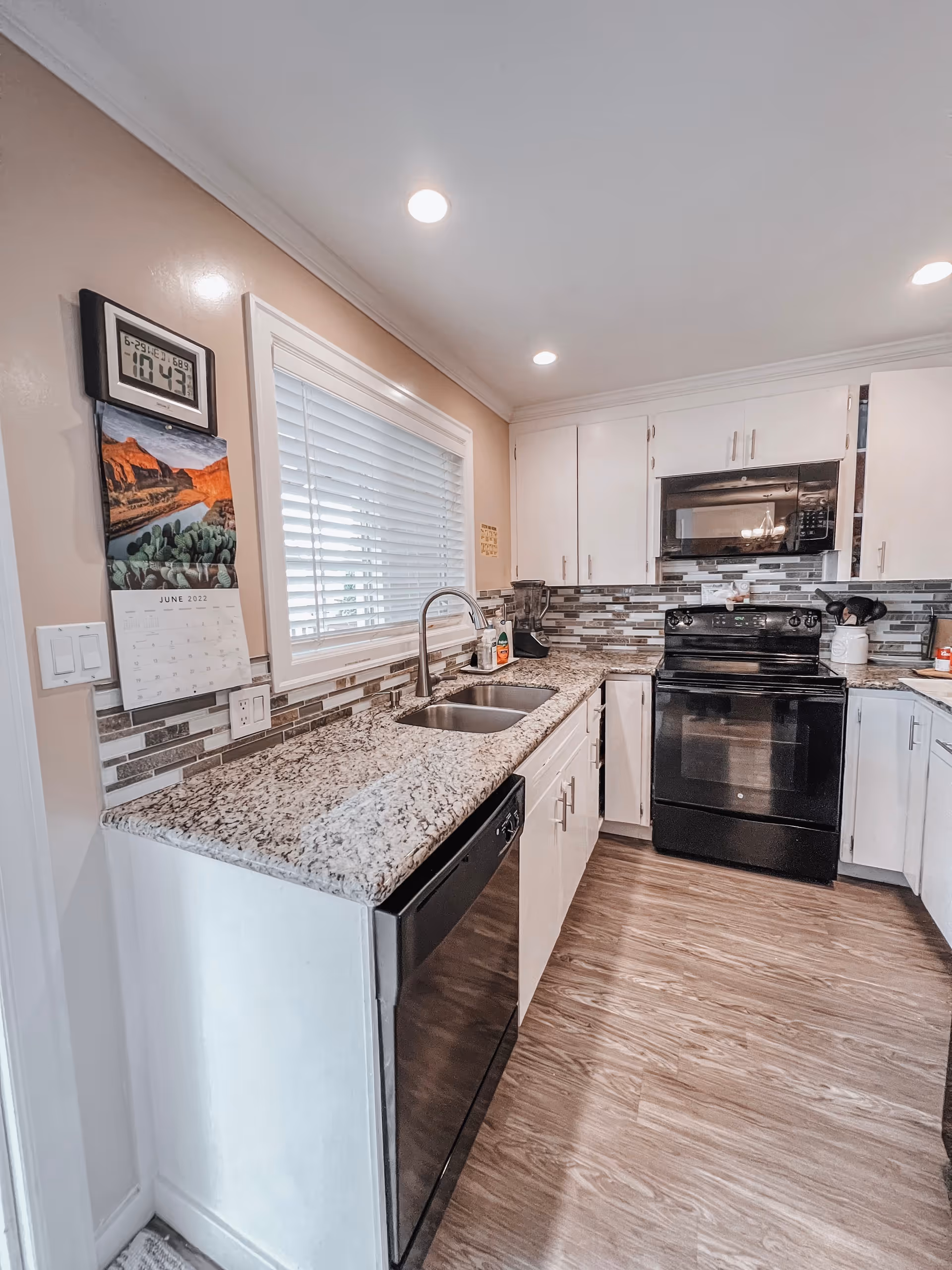 Modern kitchen with white cabinets, granite countertops, a double sink, black dishwasher, black stove, and microwave. The backsplash features a mosaic tile design in shades of gray and white. There is a window with white blinds above the sink, a calendar and digital clock on the wall, and wood-look flooring.