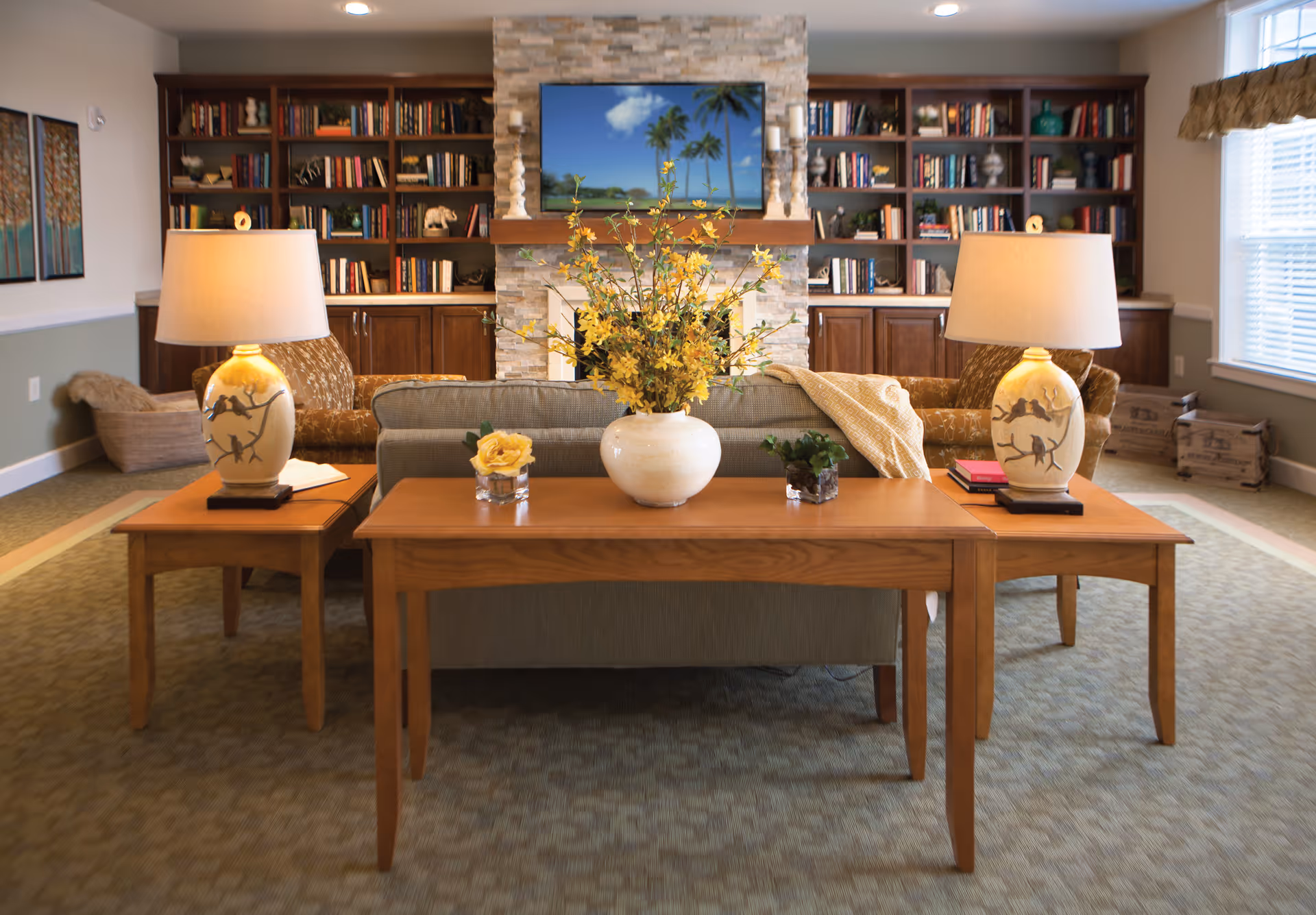 A cozy living room area with a stone fireplace and a flat-screen TV mounted above it. The room features built-in wooden bookshelves filled with books and decorative items. In front of the fireplace, there is a sofa with a wooden console table behind it, decorated with a large vase of yellow flowers and two smaller plants. Two matching wooden side tables with lamps flank the sofa. The room has carpeted flooring and large windows with blinds allowing natural light to enter.