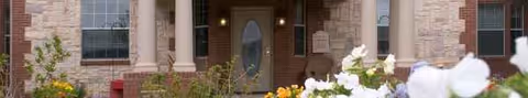 Stone-front entrance with columns, glass door, and blooming flowers along the path.