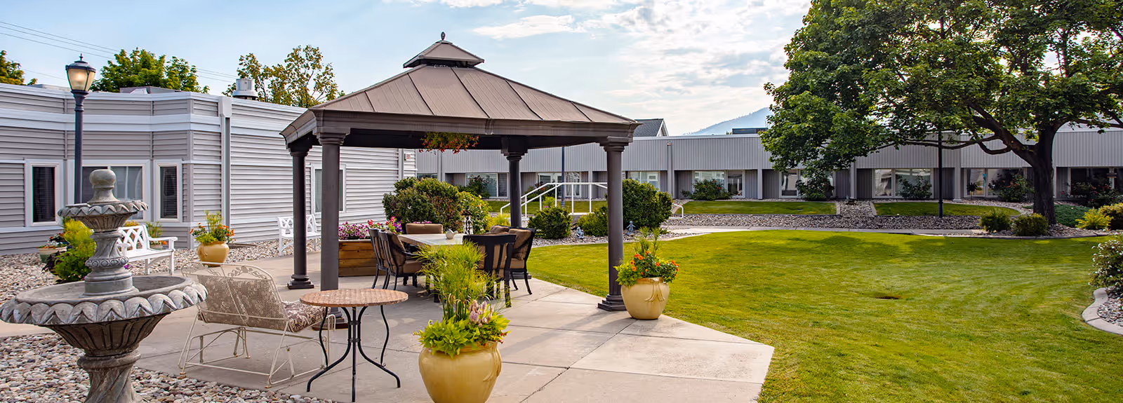 Outdoor courtyard area at The Village Health & Rehabilitation featuring a gazebo with a table and chairs underneath, surrounded by potted plants, a stone fountain, green lawn, trees, and a building with multiple windows in the background.
