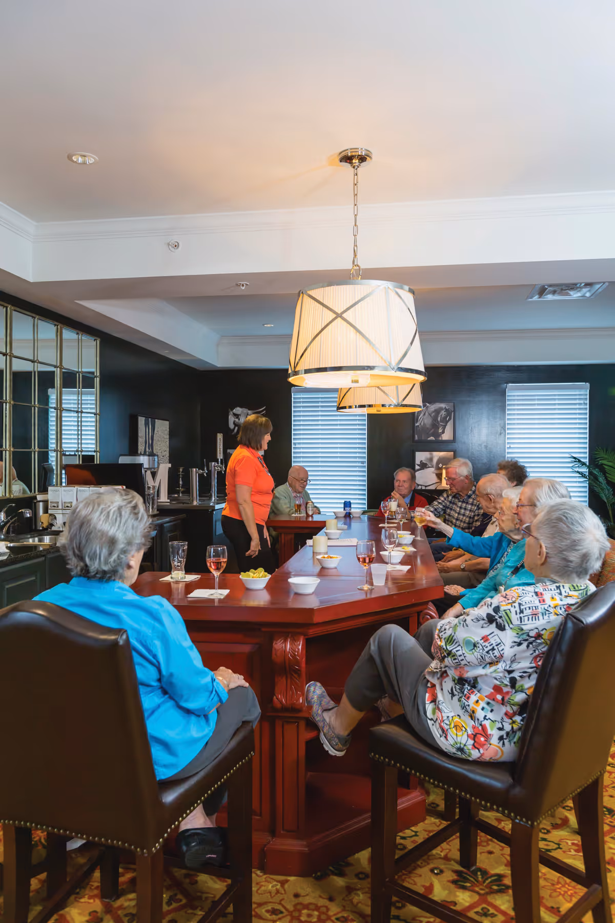 A group of elderly people sitting around a large wooden table in a well-lit room with dark walls and two windows with blinds. A woman in an orange shirt is standing and interacting with the group. There are glasses with drinks and small bowls on the table. The room has two large pendant lights hanging from the ceiling and a patterned carpet on the floor.