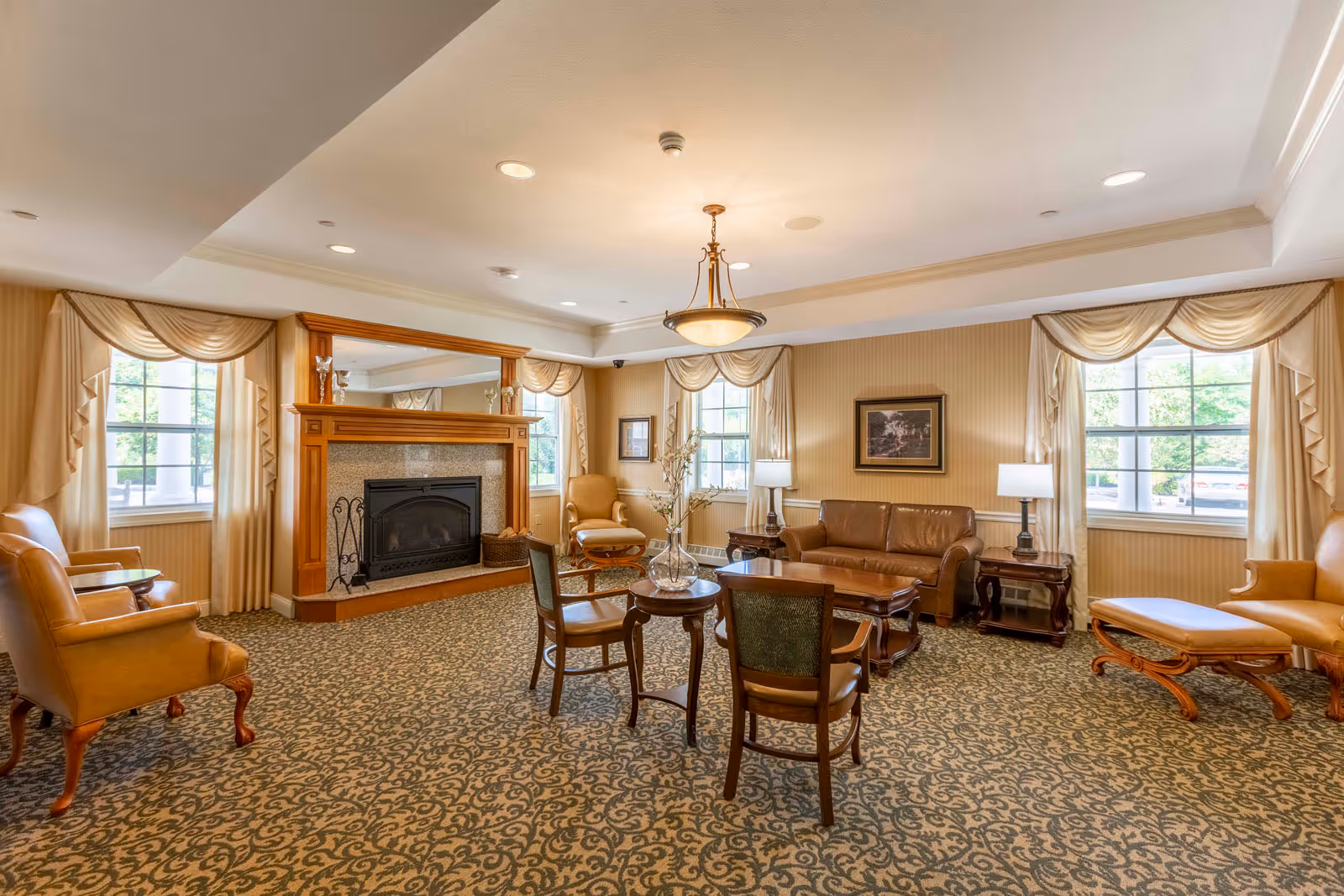 A spacious and elegant living room with patterned carpet, beige walls, and large windows with cream-colored drapes. The room features a wooden fireplace with a mirror above it, a brown leather sofa, several armchairs, a wooden coffee table, side tables with lamps, and a chandelier hanging from the ceiling. A vase with flowers is placed on a small round table in the center.