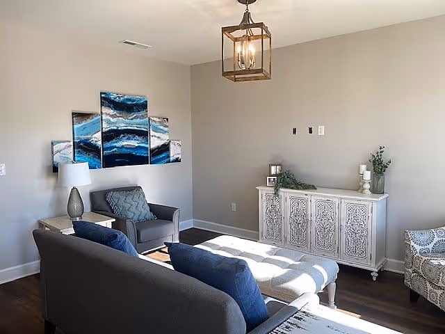 A cozy living room with a gray sofa and armchair, a white tufted ottoman, a decorative white cabinet with plants and candles on top, and a modern chandelier. Blue and white abstract artwork is displayed on the wall behind the armchair.