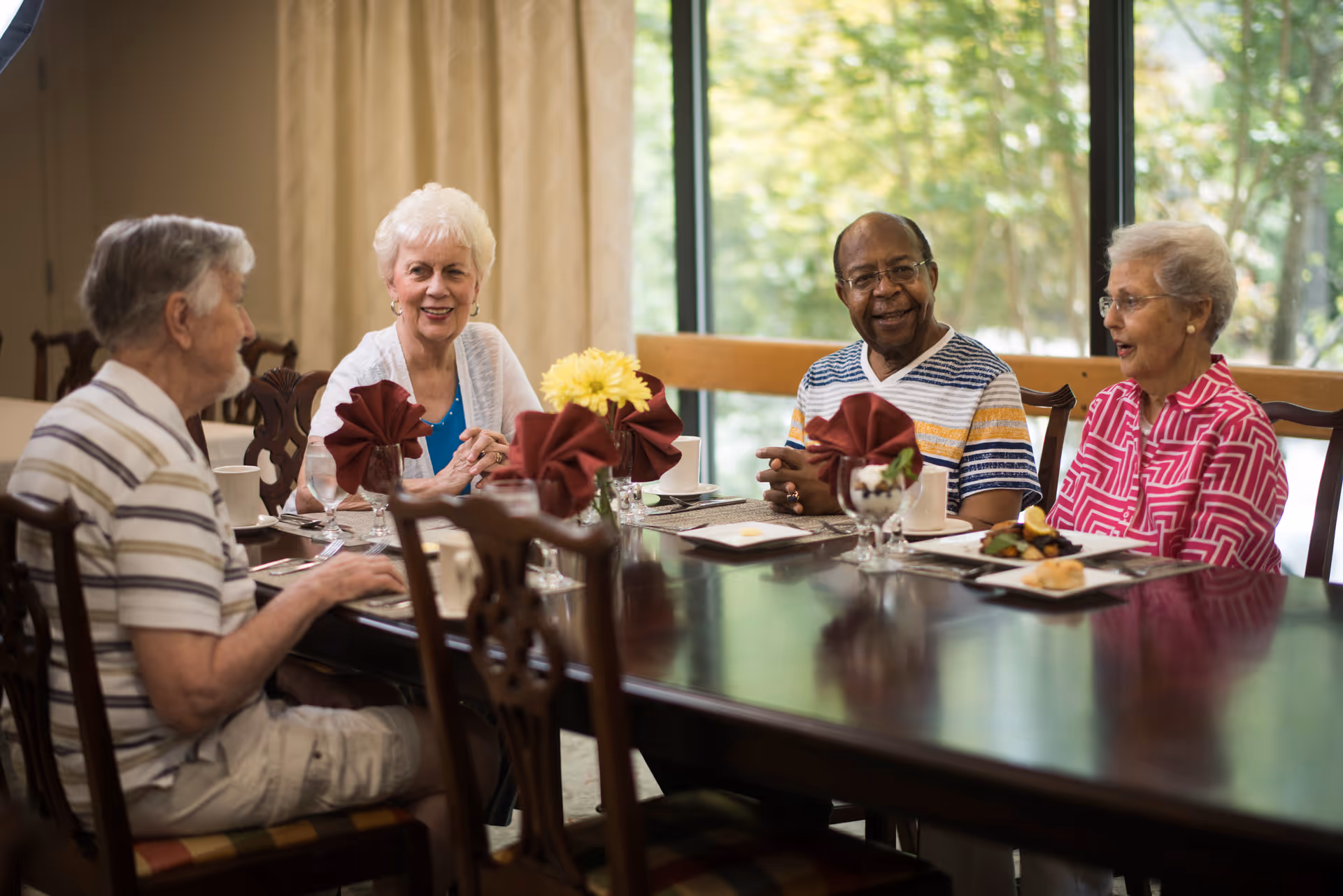 Four elderly adults seated around a dining table in a bright dining room, talking and enjoying a meal.