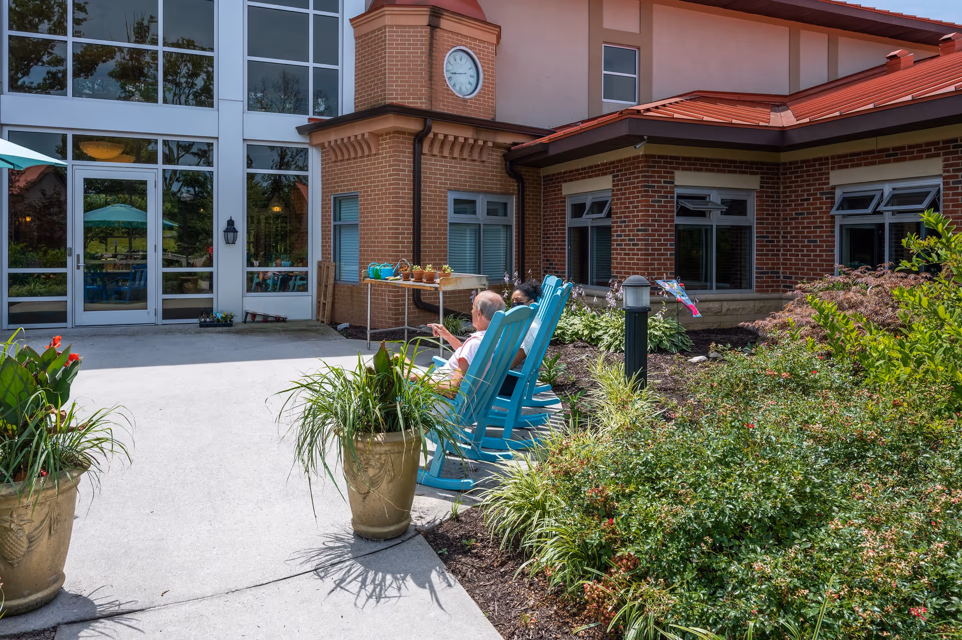 Two elderly people sitting and relaxing on blue rocking chairs outside a brick building with large windows and a clock on the wall, surrounded by plants and greenery.