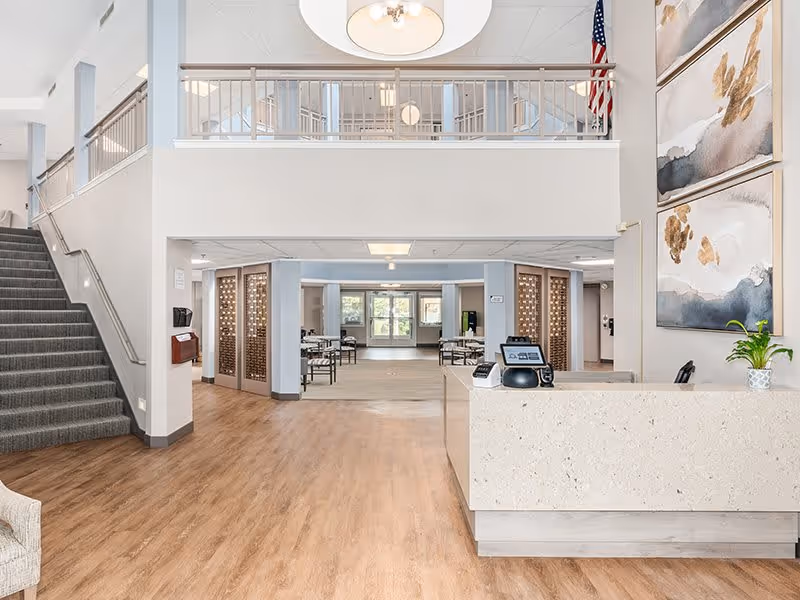 Bright and spacious senior living facility lobby with a reception desk on the right, modern light fixture hanging from the ceiling, staircase on the left, and seating area with tables and chairs in the background. Large abstract paintings and an American flag are visible on the walls.