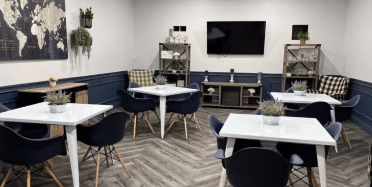 A modern common area with four white square tables each surrounded by black chairs. Each table has a small potted plant centerpiece. The room has dark blue wainscoting on the walls, light gray upper walls, and a herringbone patterned floor. There is a large flat-screen TV mounted on the far wall, flanked by two shelving units with decorative items and plants. A world map artwork and hanging plants are visible on the left wall.