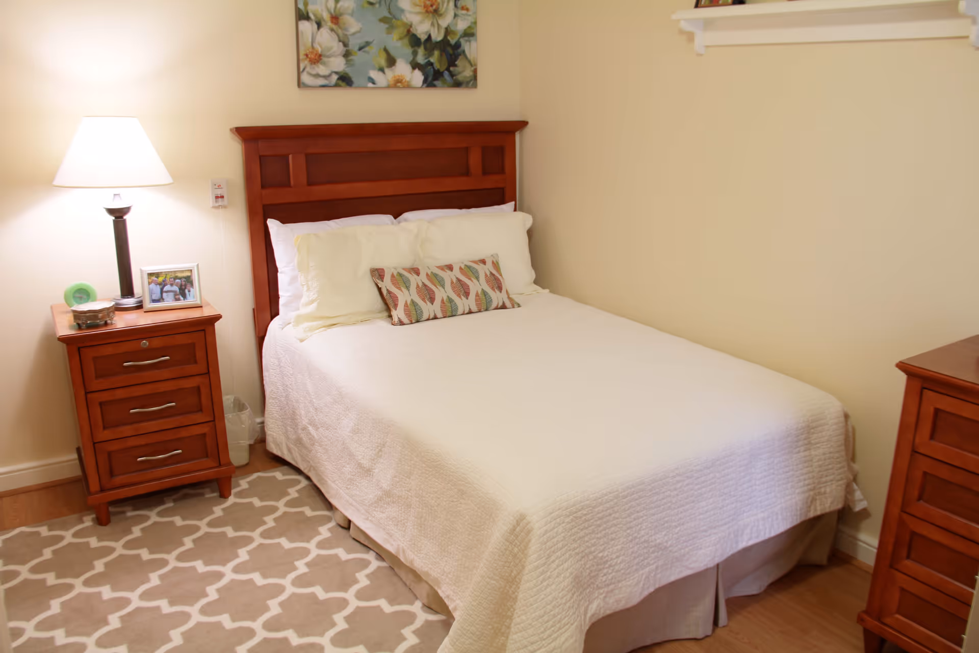 A cozy bedroom with a wooden bed frame and a neatly made bed with white bedding and decorative pillows. There is a wooden nightstand with a lamp, a framed photo, and a small decorative item. A patterned rug covers part of the wooden floor, and a floral painting hangs on the wall above the bed.