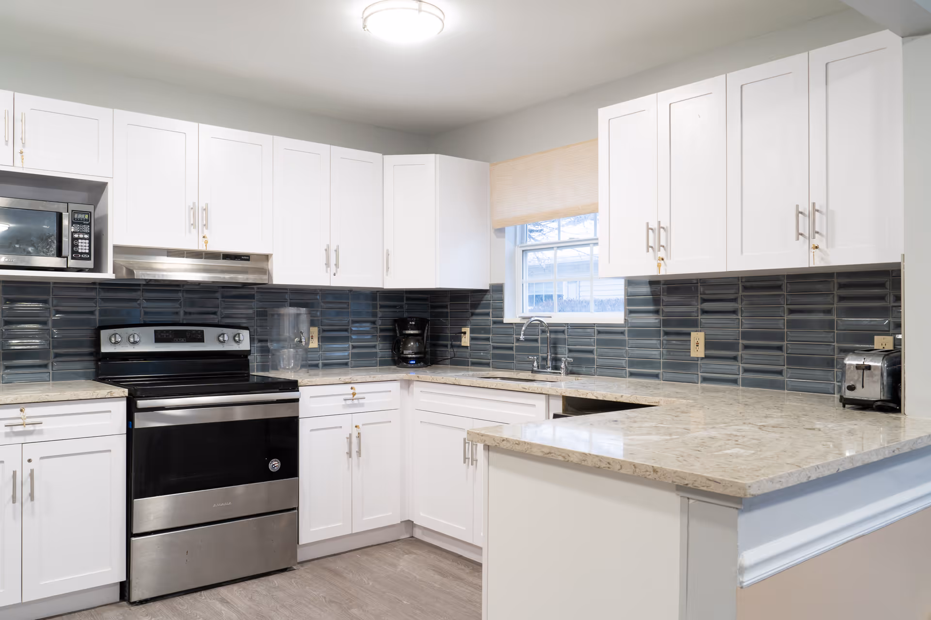 A modern kitchen with white cabinets, a stainless steel stove and microwave, a coffee maker, a toaster, and a sink under a window. The backsplash features dark blue rectangular tiles and the countertops are light-colored granite.
