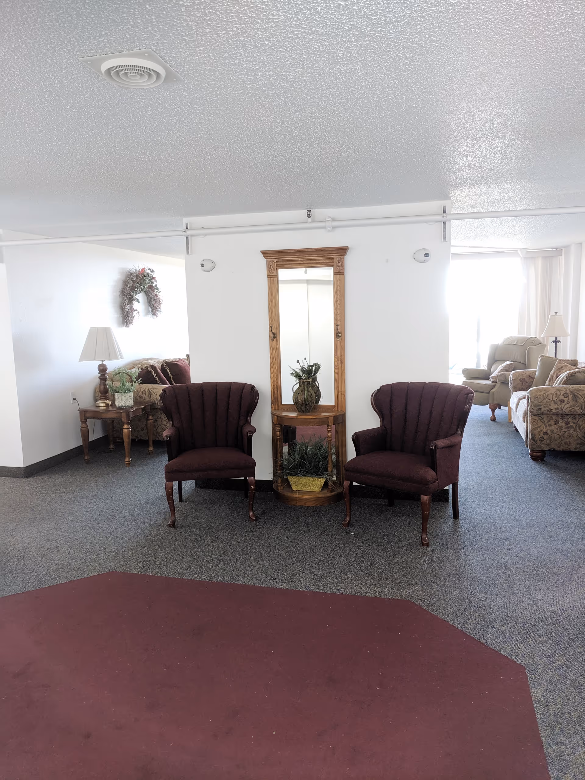 Interior view of a senior living facility lounge area with two maroon upholstered chairs flanking a wooden table with a mirror and decorative plants. In the background, there are additional seating areas with sofas and armchairs, a side table with a lamp, and a wall decoration. The floor is carpeted with a maroon and gray pattern.