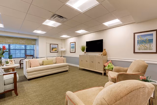 Well-lit communal living room with sofas, armchairs, a wall-mounted TV, and framed artwork.
