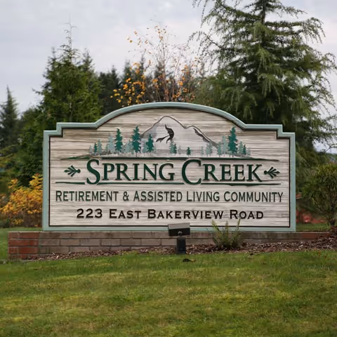 A large wooden sign for Spring Creek Retirement & Assisted Living Community at 223 East Bakerview Road, set on a brick base with green grass and trees in the background.