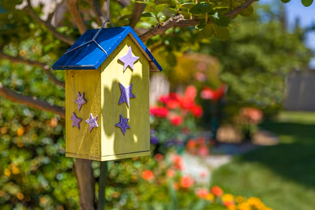 A yellow birdhouse decorated with purple stars and a blue roof hangs from a tree with a colorful garden blurred in the background.