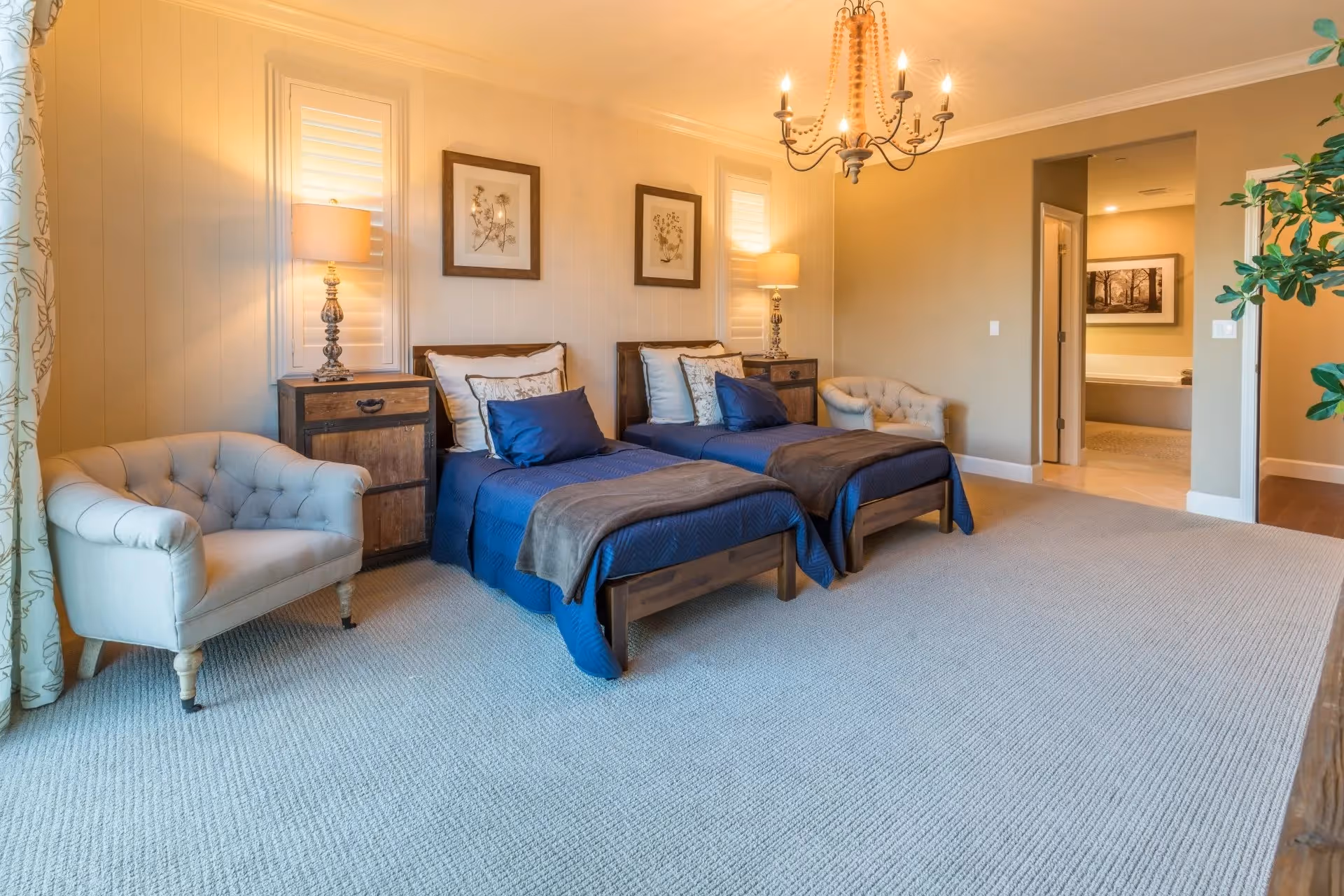 A spacious bedroom with two twin beds dressed in blue bedding and brown throws, each with multiple pillows. Between the beds is a wooden nightstand with a decorative lamp. On either side of the beds are beige tufted armchairs. The room has beige walls, carpeted floor, and two framed botanical prints above the beds. A chandelier hangs from the ceiling. An open doorway leads to a bathroom area with a bathtub and a framed picture on the wall. A green plant is partially visible on the right side of the image.