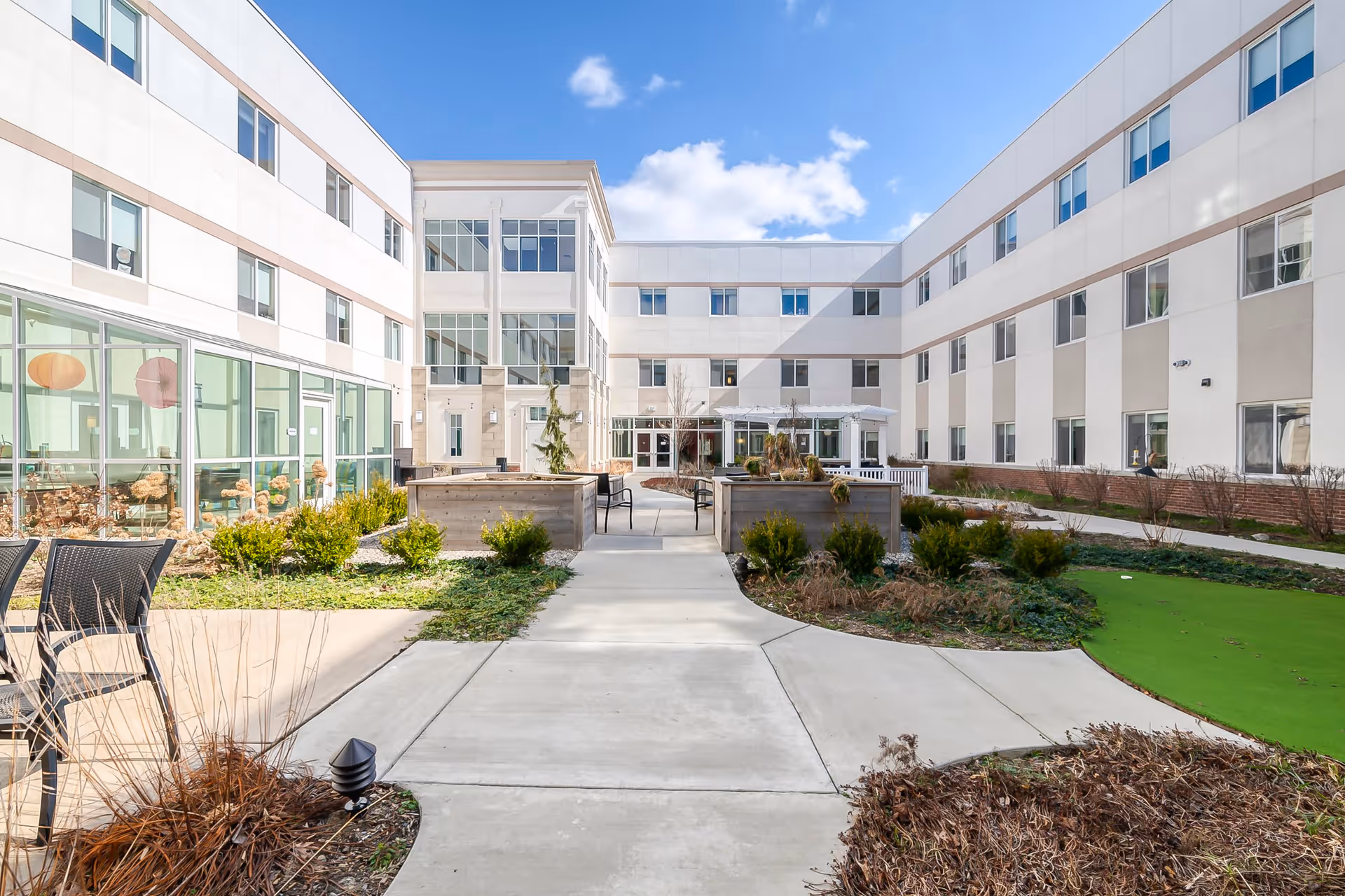 Outdoor courtyard area of The Avalon of Commerce Township senior living facility with paved walkways, green landscaping, seating areas, and a three-story building surrounding the courtyard under a blue sky with some clouds.
