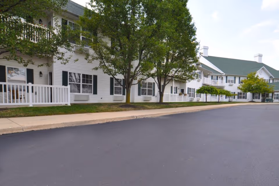 Exterior view of Jefferson Manor senior living facility showing a two-story white building with green shutters, several trees, a paved driveway, and a sidewalk.