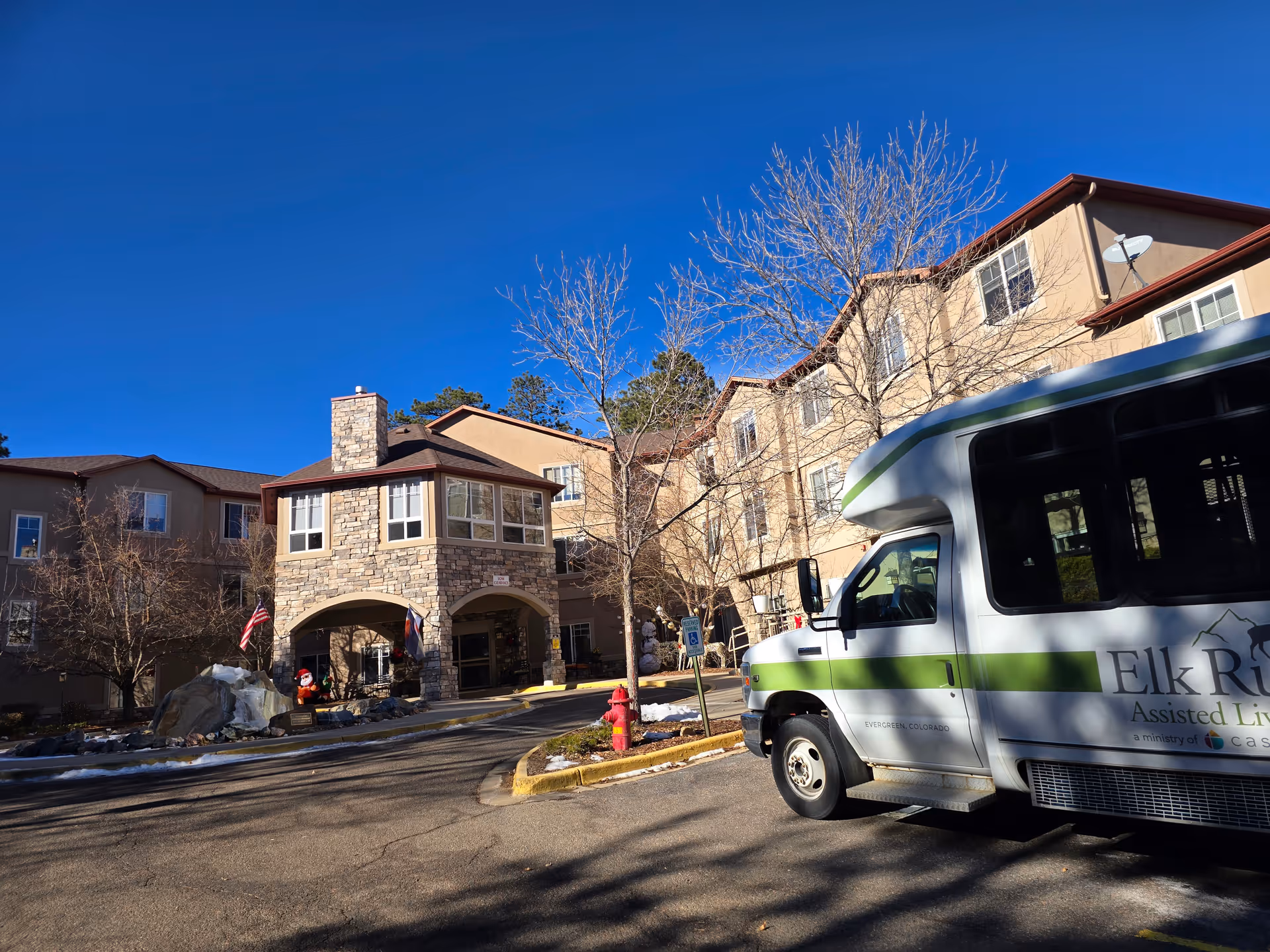 Exterior view of Elk Run Assisted Living facility showing a multi-story building with stone and beige walls, leafless trees, a clear blue sky, and a white shuttle bus with green stripes parked in front.