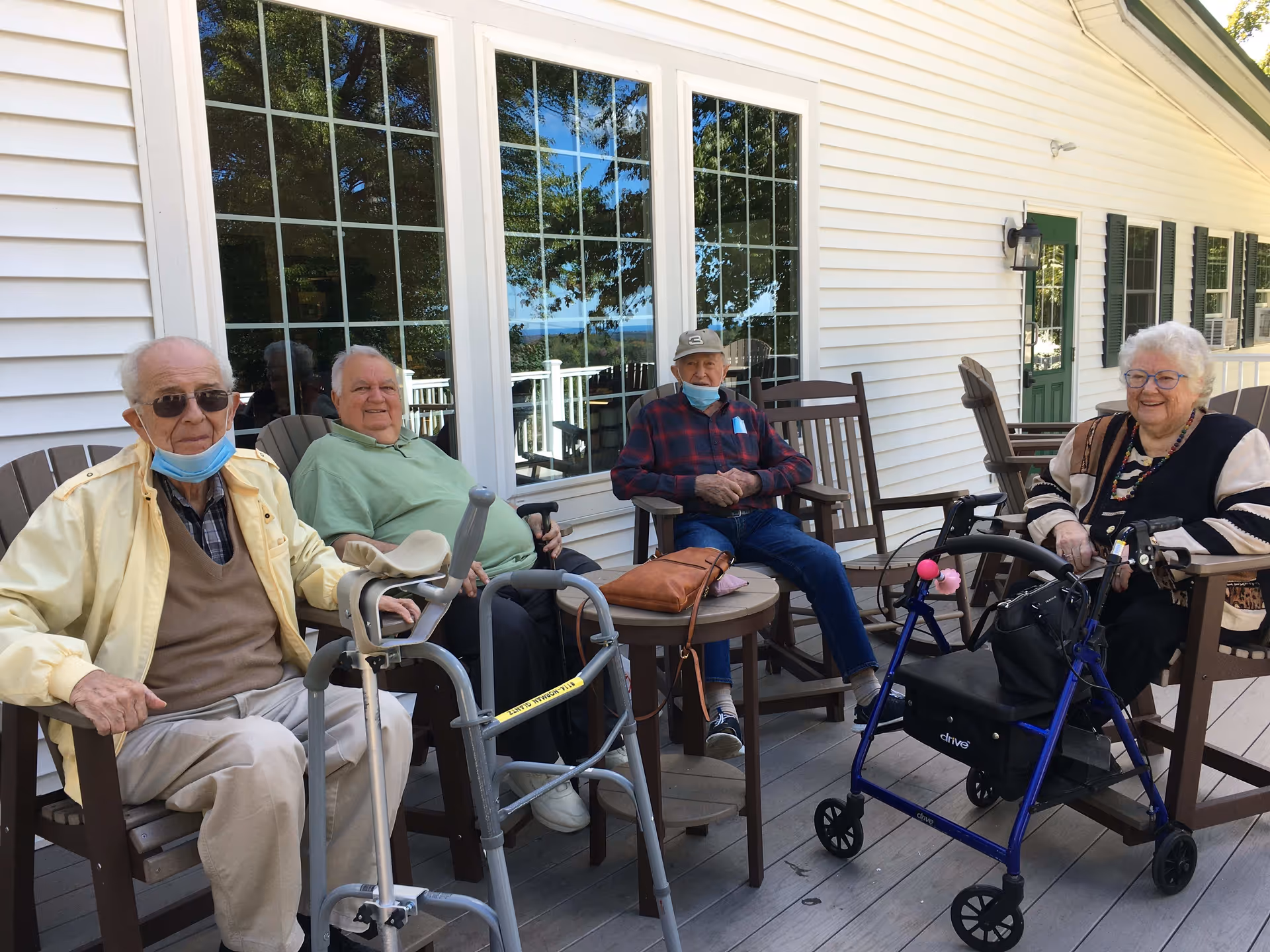 Four elderly residents sitting on a shaded outdoor deck in front of a white building, with walkers and a rolling walker nearby.