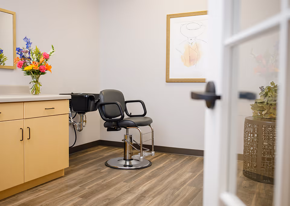 Interior view of a small salon room with a black salon chair in front of a black sink. There is a wooden cabinet with a vase of colorful flowers on the countertop. A framed artwork of a person wearing a hat hangs on the wall. The floor is wood, and a glass door with a black handle is partially open.