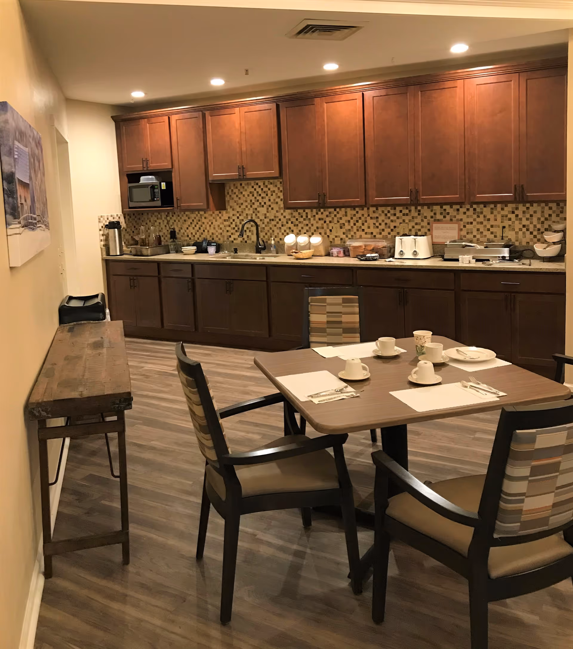 A kitchen area with wooden cabinets and a mosaic tile backsplash. There is a countertop with various kitchen appliances including a microwave, toaster, and coffee maker. In the foreground, there is a dining table set with four chairs, each with a cup and saucer, napkins, and utensils. The floor is wood-patterned, and there is a wooden console table along the left wall with a framed picture above it.