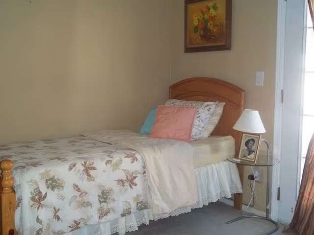A single bed with a wooden headboard in a bedroom. The bed is made with floral patterned bedding, a white blanket, and several pillows including a pink one. Next to the bed is a small round glass side table with a white lamp and a framed black and white photograph. A painting with flowers hangs on the wall above the bed. A door with glass panels is partially visible on the right side.