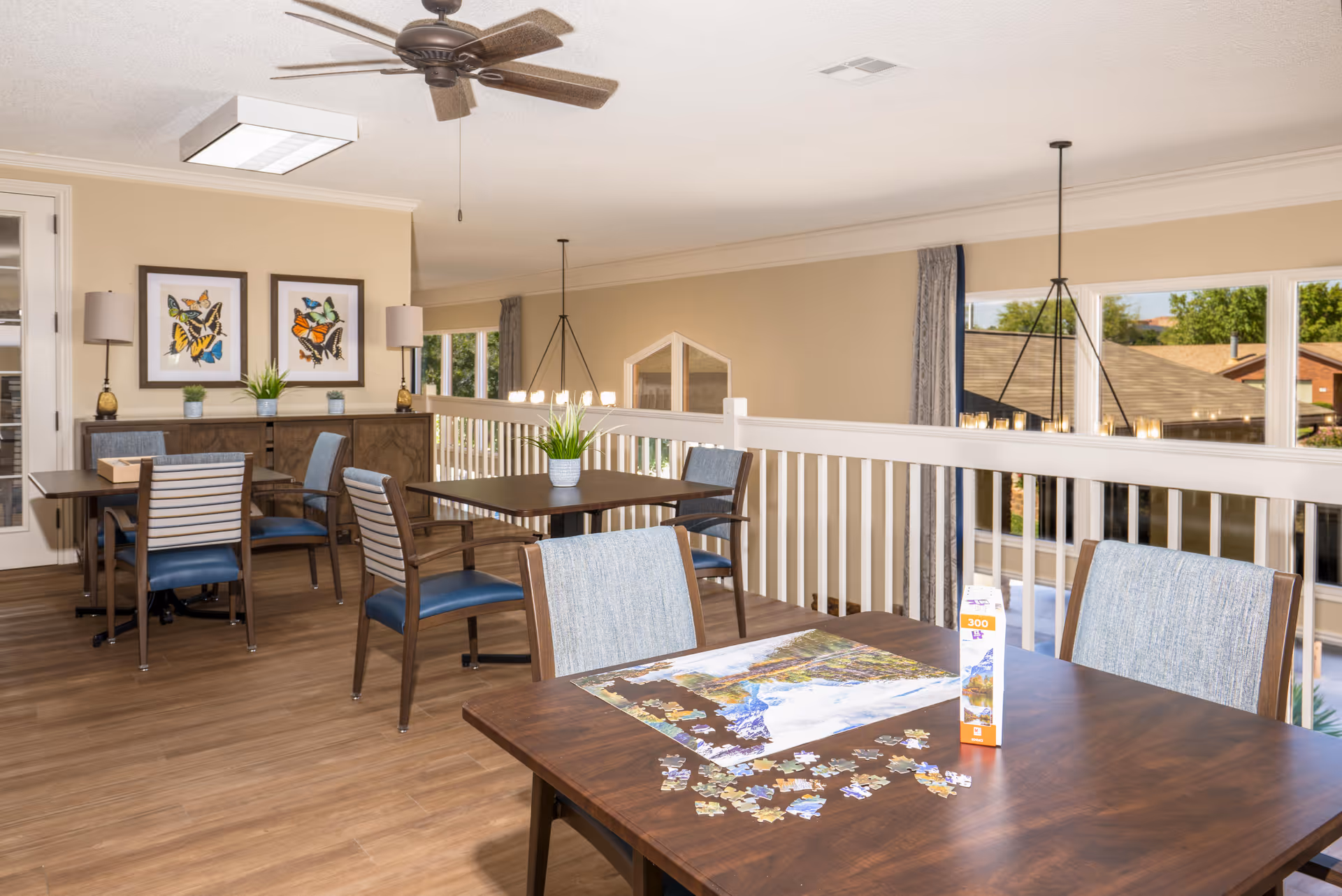 A bright and spacious common area with several wooden tables and chairs. One table in the foreground has a partially completed jigsaw puzzle and a puzzle box on it. The room features wood flooring, beige walls, large windows with views of rooftops and greenery outside, and ceiling fans. There are framed butterfly artworks on the wall and small potted plants on the tables and sideboard.