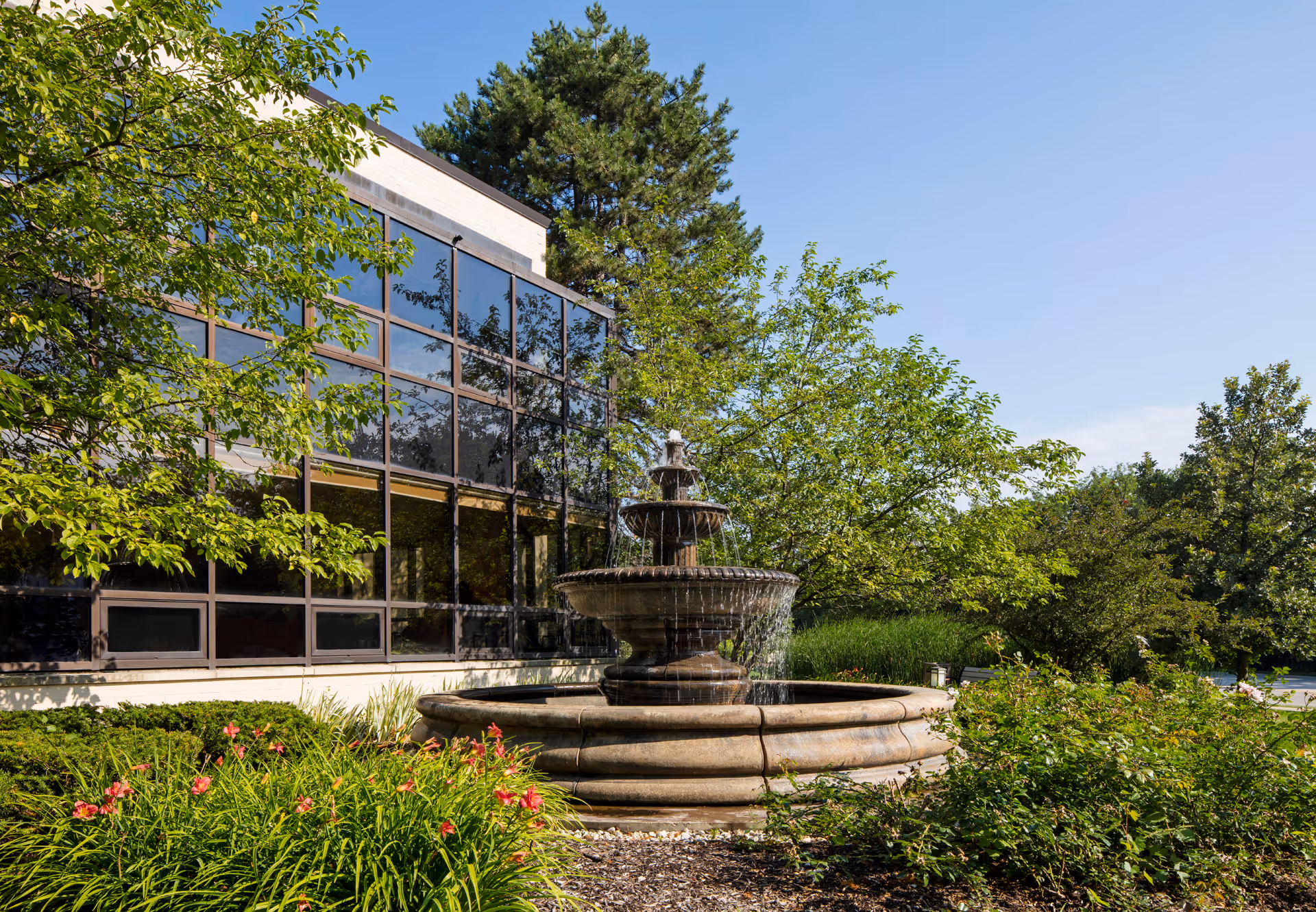 Stone tiered fountain in front of a glass-sided building surrounded by trees and flowering shrubs.