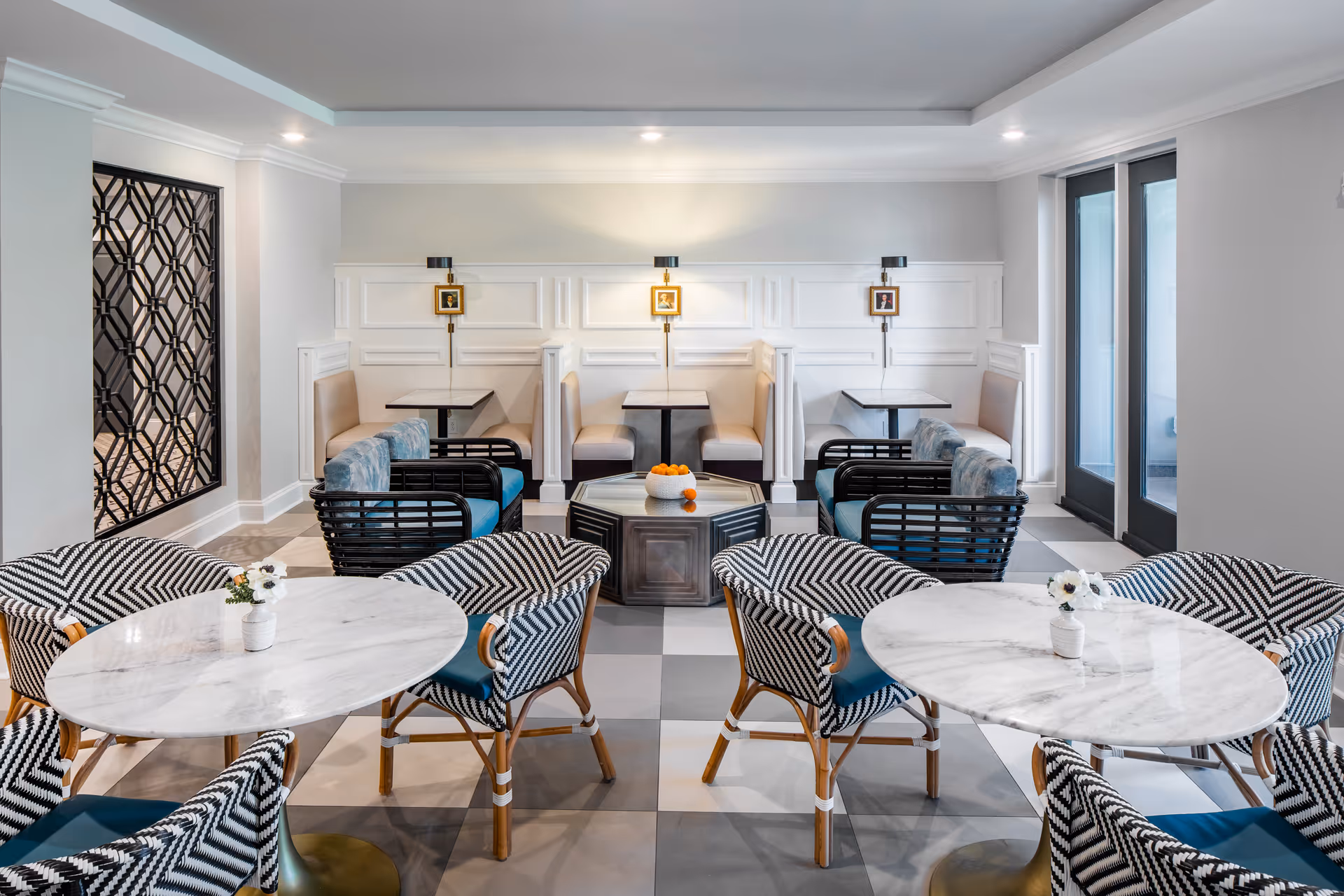 A bright and modern dining area with round marble tables surrounded by patterned chairs in the foreground. In the background, there are booth seating areas with beige cushions and small square tables. The room features a checkered tile floor, white paneled walls, and black-framed glass doors on the right side.