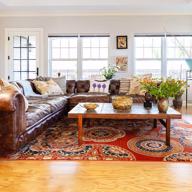 A bright living room with a large brown leather sectional sofa adorned with various pillows, a wooden coffee table with decorative bowls and vases, a colorful patterned rug, and large windows letting in natural light.