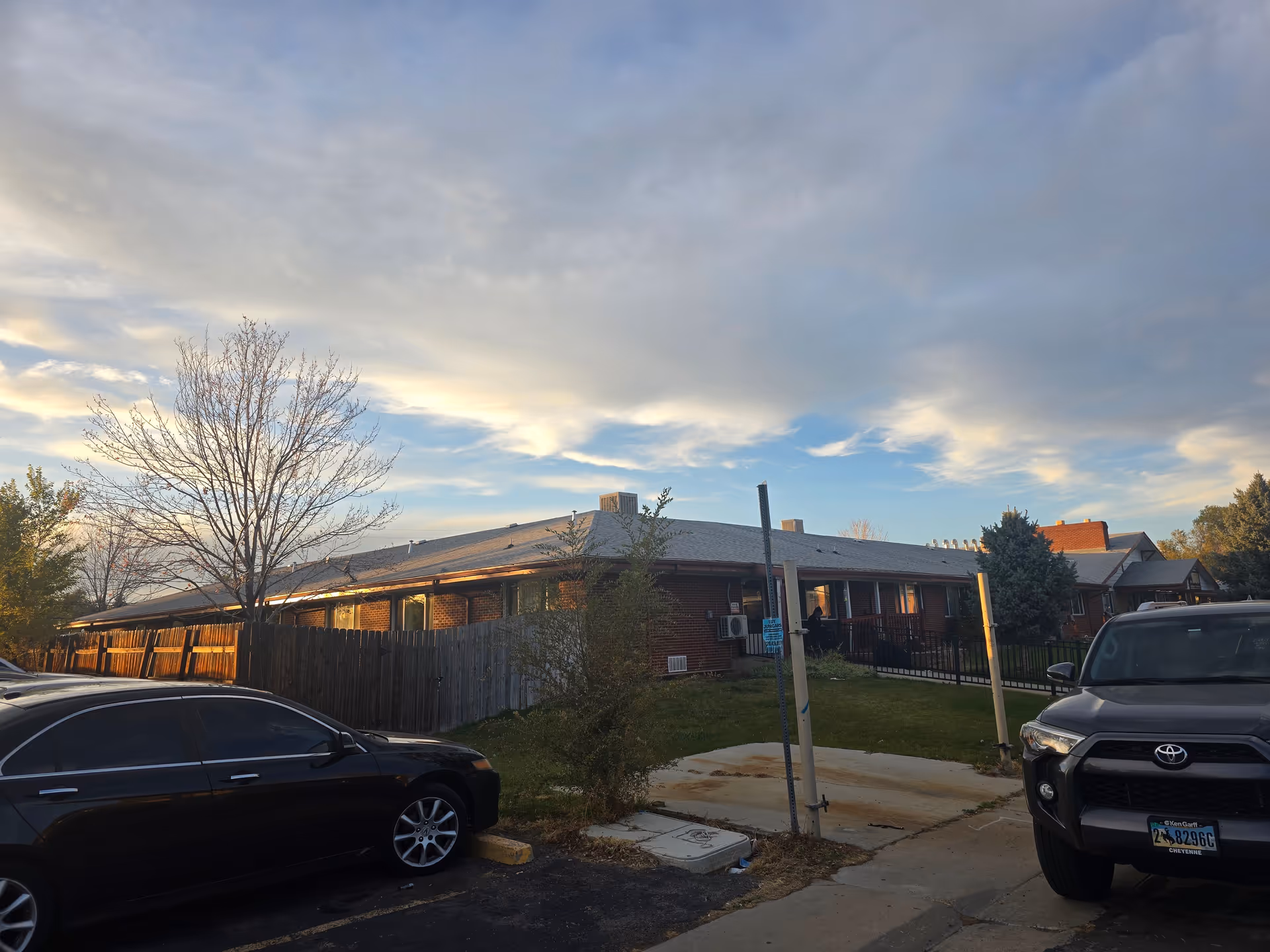 Exterior view of a single-story brick building with a fenced yard and several trees, parked cars in the foreground, under a partly cloudy sky during sunset.