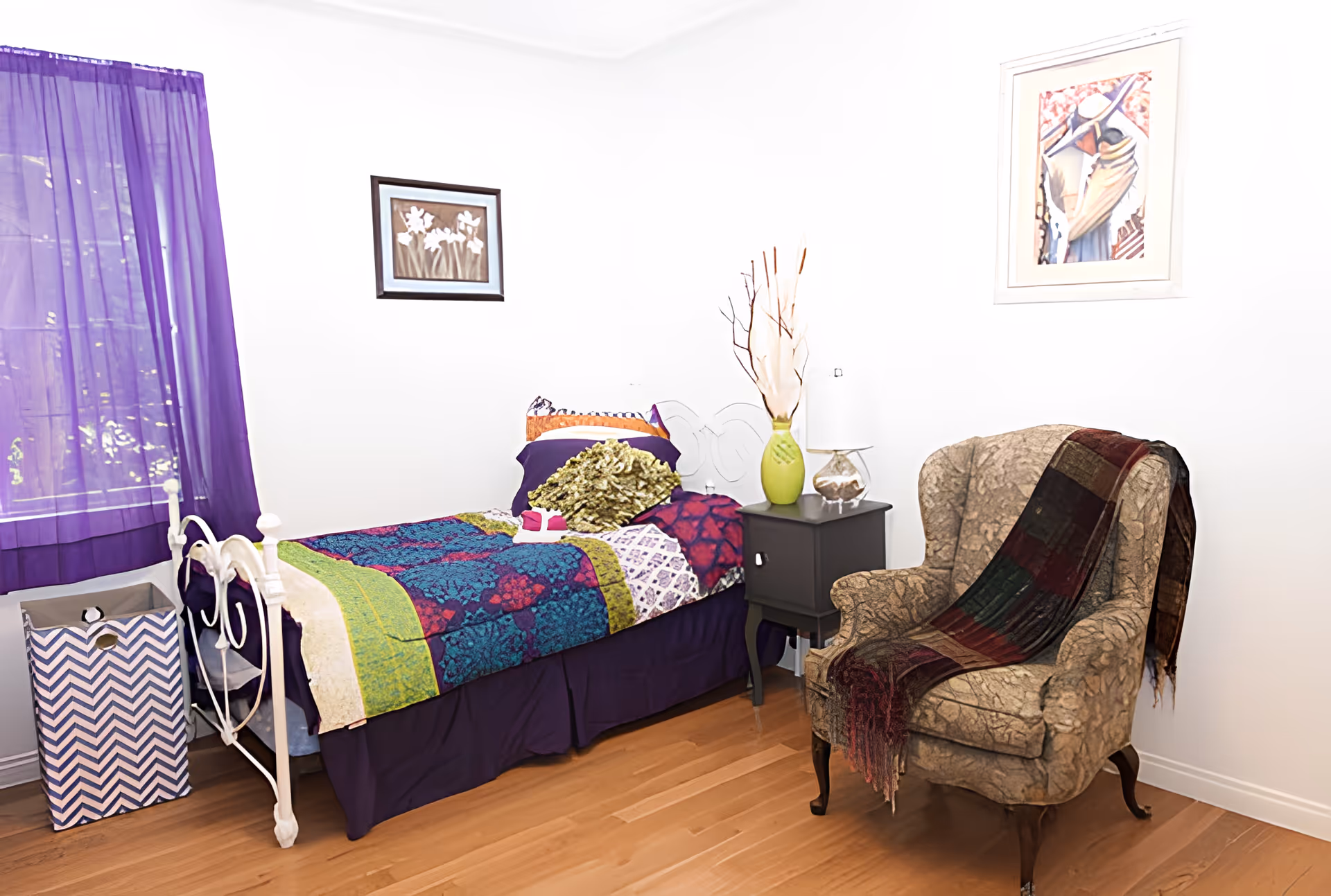 A cozy bedroom featuring a bed with colorful bedding, an armchair, and decorative elements.